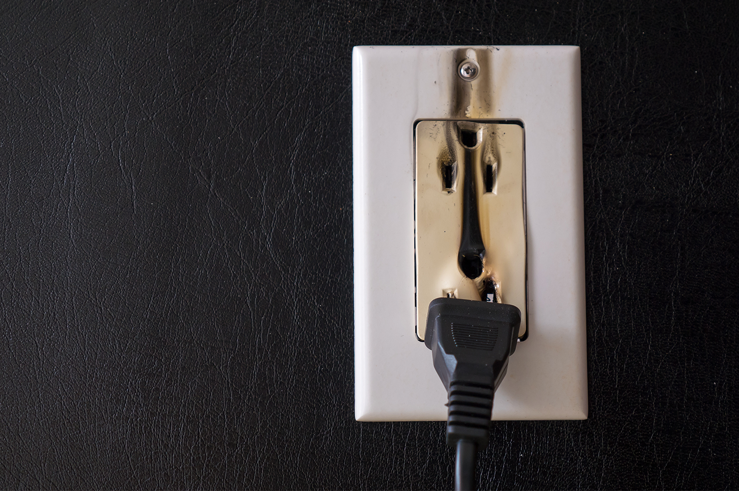 A white outlet on a black wall, with a black cord coming out of it. Black smoke is billowing from the outlet holes, indicating there's an electrical fire